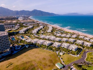 The scene captures a coastal resort area with numerous white buildings arranged in neat rows parallel to the coastline. The buildings are surrounded by lush greenery and well-maintained gardens. The turquoise ocean stretches alongside the sandy beach, while mountains rise in the background under a clear blue sky.