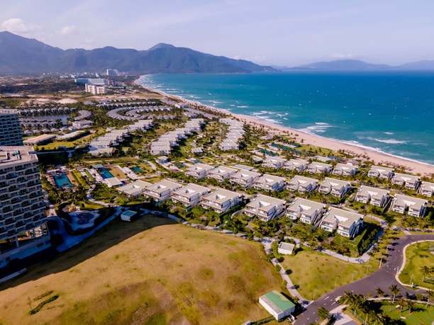 The scene captures a coastal resort area with numerous white buildings arranged in neat rows parallel to the coastline. The buildings are surrounded by lush greenery and well-maintained gardens. The turquoise ocean stretches alongside the sandy beach, while mountains rise in the background under a clear blue sky.