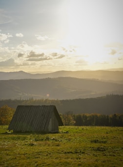 A solitary wooden hut stands on a grassy field, with a serene backdrop of rolling hills and a partially cloudy sky illuminated by the warm glow of sunset. The landscape creates a peaceful and idyllic rural scene.