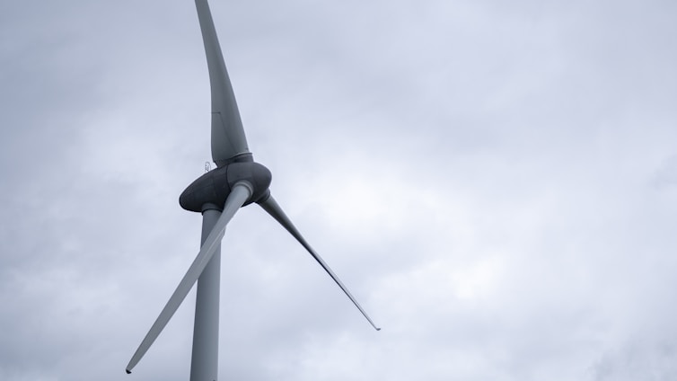 A large wind turbine with three blades set against a cloudy sky. The structure appears sturdy and industrial, with smooth, aerodynamic blades extending from a central hub.
