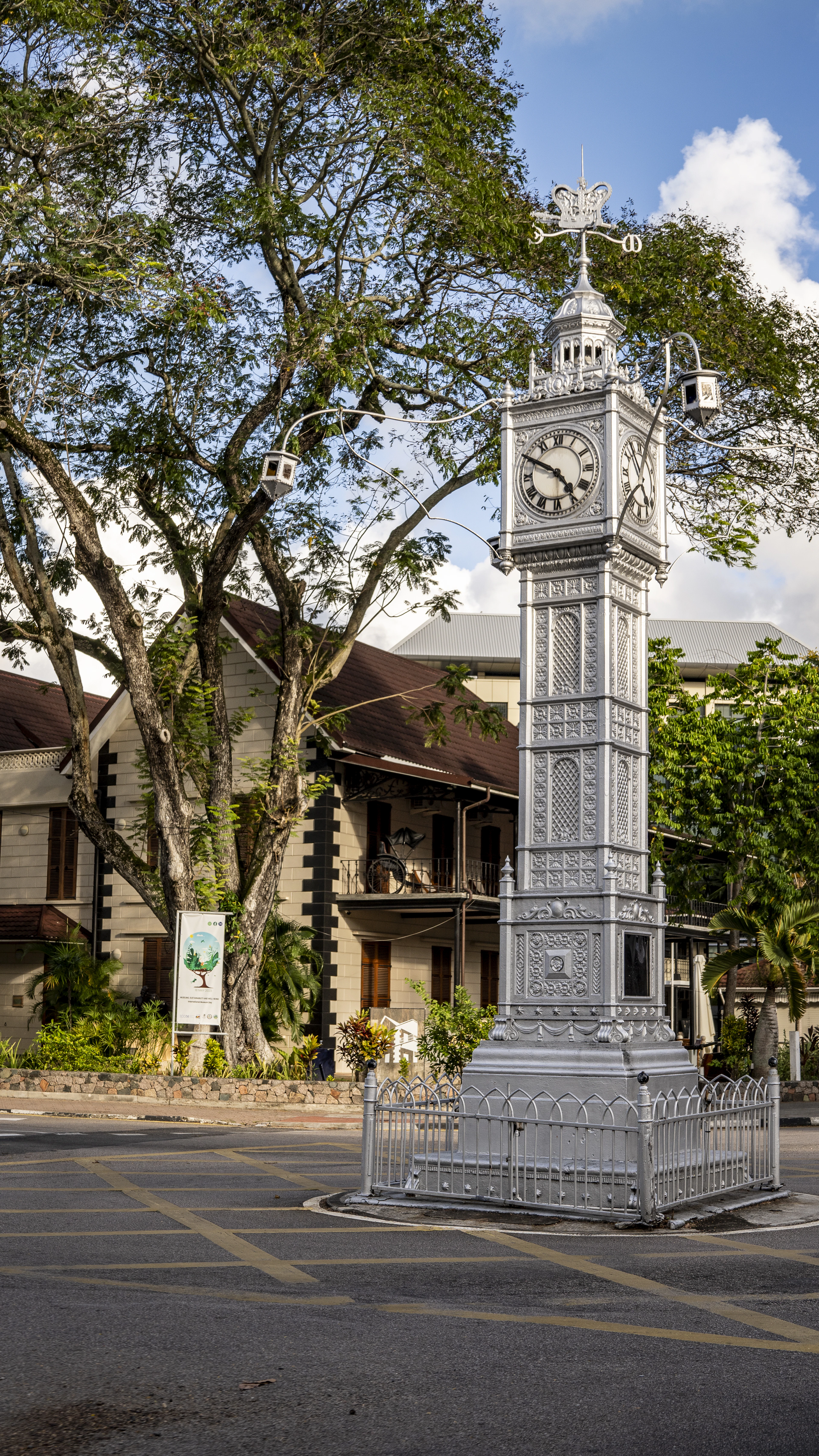 The silver cast-iron Victoria clocktower stands on its little road island in the centre of Victoria, Mahe, backed by a colonial verandah building and a broad shade tree