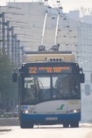 A trolleybus is traveling on an urban street, featuring an overhead power supply with wires connecting to the bus's roof. The front display shows the route number and destination in bright letters. The environment includes buildings and infrastructure typical of a city setting.