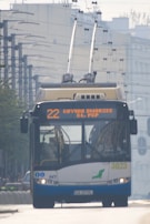 A trolleybus is traveling on an urban street, featuring an overhead power supply with wires connecting to the bus's roof. The front display shows the route number and destination in bright letters. The environment includes buildings and infrastructure typical of a city setting.