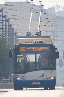 A trolleybus is traveling on an urban street, featuring an overhead power supply with wires connecting to the bus's roof. The front display shows the route number and destination in bright letters. The environment includes buildings and infrastructure typical of a city setting.