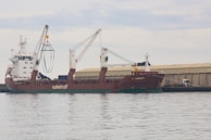A large cargo ship is docked at a port with two cranes extending from the vessel. The ship is named 'Spliethoff' and 'Faglegracht', with signage visible on the side. A warehouse is situated in the background, and a truck is parked nearby. The sky is overcast, reflecting off the calm water surface.