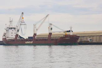 A large cargo ship is docked at a port with two cranes extending from the vessel. The ship is named 'Spliethoff' and 'Faglegracht', with signage visible on the side. A warehouse is situated in the background, and a truck is parked nearby. The sky is overcast, reflecting off the calm water surface.