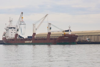A large cargo ship is docked at a port with two cranes extending from the vessel. The ship is named 'Spliethoff' and 'Faglegracht', with signage visible on the side. A warehouse is situated in the background, and a truck is parked nearby. The sky is overcast, reflecting off the calm water surface.