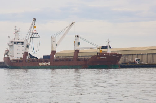 A large cargo ship is docked at a port with two cranes extending from the vessel. The ship is named 'Spliethoff' and 'Faglegracht', with signage visible on the side. A warehouse is situated in the background, and a truck is parked nearby. The sky is overcast, reflecting off the calm water surface.