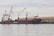A large cargo ship is docked at a port with two cranes extending from the vessel. The ship is named 'Spliethoff' and 'Faglegracht', with signage visible on the side. A warehouse is situated in the background, and a truck is parked nearby. The sky is overcast, reflecting off the calm water surface.