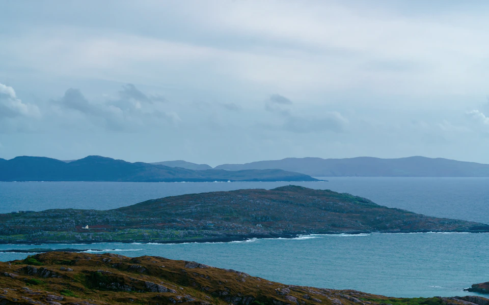 The image depicts a serene coastal landscape with a large, light blue body of water stretching out towards the horizon. The water is dotted with small islands scattered across its surface, adding depth and texture to the scene. In the foreground, there are several rocky hills that rise up from the water's edge, their dark brown color contrasting with the lighter tones of the surrounding landscape.

The sky above is a light blue canvas, lightly brushed with wispy clouds that add movement and dynamism to the otherwise tranquil setting. The image is taken from an elevated perspective, offering a panoramic view of the water's edge and the distant hills.