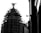 A black and white image features a building under construction or renovation, covered with scaffolding. Atop the structure is a silhouetted statue of a winged figure. The right side of the image shows part of a wall with some architectural details. The overall contrast creates a dramatic effect.