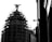 A black and white image features a building under construction or renovation, covered with scaffolding. Atop the structure is a silhouetted statue of a winged figure. The right side of the image shows part of a wall with some architectural details. The overall contrast creates a dramatic effect.