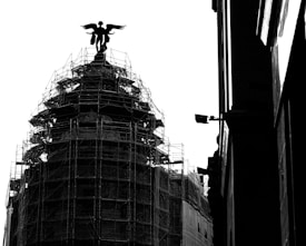 A black and white image features a building under construction or renovation, covered with scaffolding. Atop the structure is a silhouetted statue of a winged figure. The right side of the image shows part of a wall with some architectural details. The overall contrast creates a dramatic effect.