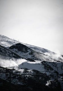A snowy mountain landscape with ski lifts ascending the slopes. Snow covers the ground, with some rocky patches visible. Snow cannons are in operation, emitting plumes of snow. In the foreground, several gondola lift cars can be seen.