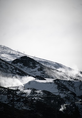 A snowy mountain landscape with ski lifts ascending the slopes. Snow covers the ground, with some rocky patches visible. Snow cannons are in operation, emitting plumes of snow. In the foreground, several gondola lift cars can be seen.