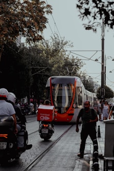 A modern tram navigates through a busy urban street lined with trees. People are walking on the sidewalks, and a person is riding a scooter near the tram. There are hints of greenery and street lamps on the sides.