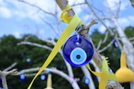 A blue Nazar amulet hangs from a tree branch, tied with a bright yellow ribbon. The background consists of out-of-focus green foliage and a clear blue sky with scattered clouds. Additional similar amulets and colored ornaments are visible in the distance.