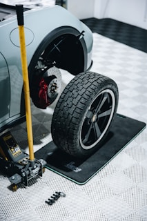 A car raised on a jack with a removed wheel sitting nearby. The car's brake system, including a red brake caliper, is visible. A set of lug nuts and the yellow car jack are placed on a checkered garage floor.