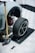 A car raised on a jack with a removed wheel sitting nearby. The car's brake system, including a red brake caliper, is visible. A set of lug nuts and the yellow car jack are placed on a checkered garage floor.