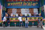 A carnival game booth with a colorful sign that reads 'Whopper Water' features rows of plush stuffed animals as prizes. Several people are standing at the counter, engaging in the game. Each workstation is equipped with a water gun. The booth is decorated with vibrant colors and patterns, including blue, green, red, and yellow.
