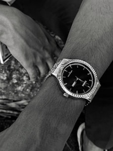A close-up view of a wrist wearing a silver wristwatch with a black clock face, showing the brand name 'Timex'. A second hand can be seen resting on a textured surface with a visible hand and a pair of legs in the background.