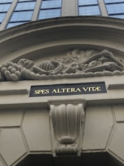 A detailed architectural bas-relief shows a skeleton amidst elaborate foliage, positioned above a building entrance. Below the relief is an inscription in gold lettering on a dark background reading 'SPES ALTERA VITÆ'. The surrounding structure features classic architectural elements in beige tones.