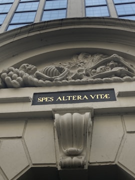 A detailed architectural bas-relief shows a skeleton amidst elaborate foliage, positioned above a building entrance. Below the relief is an inscription in gold lettering on a dark background reading 'SPES ALTERA VITÆ'. The surrounding structure features classic architectural elements in beige tones.
