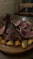A wooden plate filled with thick slices of rare-cooked beef layered on top of roasted potatoes. The cut of meat is juicy with a pink interior and a dark brown seared exterior. A wooden chair and part of a person’s leg are visible in the dim, rustic setting.