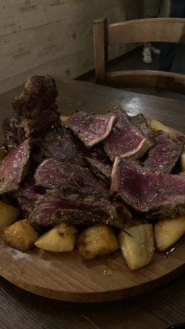 A wooden plate filled with thick slices of rare-cooked beef layered on top of roasted potatoes. The cut of meat is juicy with a pink interior and a dark brown seared exterior. A wooden chair and part of a person’s leg are visible in the dim, rustic setting.