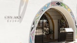An arched entrance with colorful geometric patterns frames the entryway, featuring a black metal gate. The wall is painted white with the inscription 'CAVALARICA ÉVORA' written on it. A potted plant is visible inside the entrance, and there is a modern-looking stand on the right side with a reflective surface.