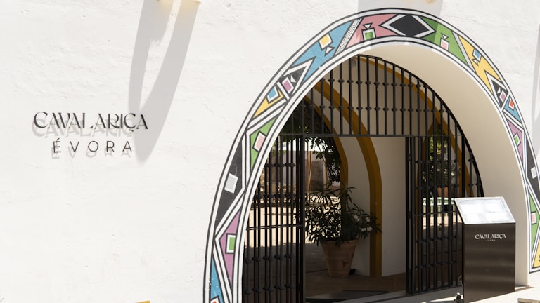 An arched entrance with colorful geometric patterns frames the entryway, featuring a black metal gate. The wall is painted white with the inscription 'CAVALARICA ÉVORA' written on it. A potted plant is visible inside the entrance, and there is a modern-looking stand on the right side with a reflective surface.