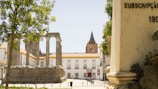 Historic Roman temple ruins with tall stone columns stand in a sunlit square surrounded by white buildings with terracotta roofs. Mature trees provide shade and a tower is visible in the background. Architectural details and a monument are also present.