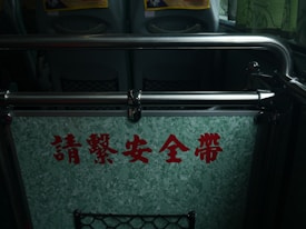 A barrier inside a vehicle, likely a bus, featuring a sign with red Chinese characters on a textured green panel. The metal railings frame the sign and seats are visible in the background, suggesting a public transportation setting. There are also pockets on the seatbacks.