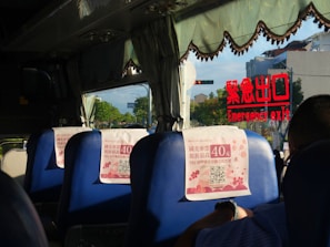 Inside a bus with blue seats, each having a promotional flyer attached. The window displays an emergency exit sign in red lettering reflected on the glass. Outside, trees and a few buildings are visible through the window.