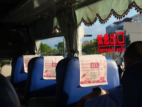 Inside a bus with blue seats, each having a promotional flyer attached. The window displays an emergency exit sign in red lettering reflected on the glass. Outside, trees and a few buildings are visible through the window.