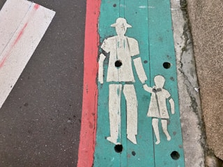 A painted pedestrian crossing symbol on a sidewalk depicting an adult holding a child's hand. The symbol is marked in white on a turquoise background, with a red line running parallel to the symbol. The sidewalk and road surface show some wear and tear.