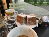 A round table holds two cubes of cake topped with powdered sugar on a white plate. Nearby, there is a coffee cup with colorful text branding, a glass of water, and a metal holder with packets of sugar. The background reveals a street view with greenery and paving.