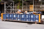 A large dumpster filled with various items, including black garbage bags, cardboard boxes, and metal chairs, is situated on a street. Scaffolding and a tree are visible behind the dumpster, which is painted blue with the word 'LIBERTY' stenciled on the side in white letters.