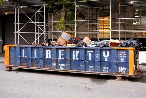 A large dumpster filled with various items, including black garbage bags, cardboard boxes, and metal chairs, is situated on a street. Scaffolding and a tree are visible behind the dumpster, which is painted blue with the word 'LIBERTY' stenciled on the side in white letters.