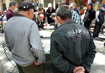 A group of older adults are gathered outdoors, closely watching a game being played on a table. The scene is in a park-like setting with trees in the background. Most people are dressed in casual jackets and caps, and some are wearing face masks.
