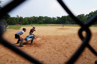 A baseball game taking place on a sandy field, viewed through a diamond-shaped fence. Several players are on the field, including a pitcher mid-throw, a batter holding a bat, a catcher crouched behind the batter, and an umpire observing closely. The scene is set against a background of lush green trees and a clear sky.
