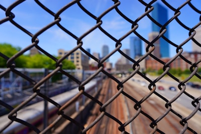 A chain-link fence is in sharp focus in the foreground, while the background shows a blurred view of railway tracks and a city skyline with several high-rise buildings. The sky is clear, indicating a sunny day.