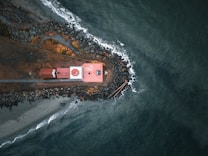 An aerial view of a small jetty extending into the sea, with a red-roofed building near the tip. Surrounding the structure are rocks and a narrow beach with driftwood scattered along the shore. Waves gently crash against the rocks, creating a dynamic contrast with the calm, deep blue of the ocean.