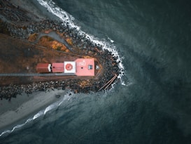 An aerial view of a small jetty extending into the sea, with a red-roofed building near the tip. Surrounding the structure are rocks and a narrow beach with driftwood scattered along the shore. Waves gently crash against the rocks, creating a dynamic contrast with the calm, deep blue of the ocean.
