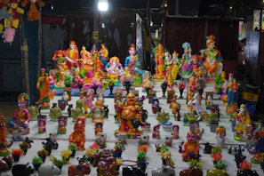 A collection of brightly colored statues and figurines is arranged on a table. The figures include various deities, animals, and mythical characters, each detailed with vibrant colors and intricate designs. The background is a dimly lit setting, emphasizing the vividness of the sculptures.