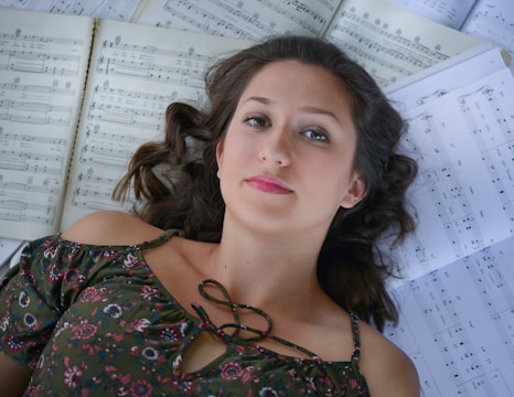 A woman with curly hair lies on her back surrounded by open sheets of music. She wears a floral patterned top with an off-shoulder design. Her expression is calm and focused.