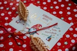 A festive holiday card with a gnome illustration, surrounded by words like 'Glück', 'Gesundheit', and 'Familie' in German. The background features red wrapping paper with white polka dots and green star designs. Decorative wooden ornaments and red string are placed on top of the envelope.