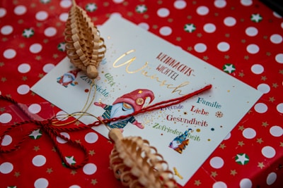 A festive holiday card with a gnome illustration, surrounded by words like 'Glück', 'Gesundheit', and 'Familie' in German. The background features red wrapping paper with white polka dots and green star designs. Decorative wooden ornaments and red string are placed on top of the envelope.