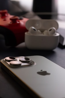 A close-up shot of a smartphone with a visible camera module and the Apple logo is placed on a dark surface. Nearby, a red gaming controller is partially visible, along with a white case containing wireless earbuds.