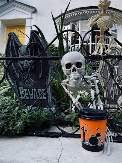 A Halloween-themed display is set in a garden with tombstones and skeletons. A realistic skull is positioned next to ironwork that spells 'Beware'. A humorous touch is added by a skeleton figure holding an orange coffee cup featuring Halloween illustrations, including a skeleton and bats. The backdrop is a classic white house with a yellow door.